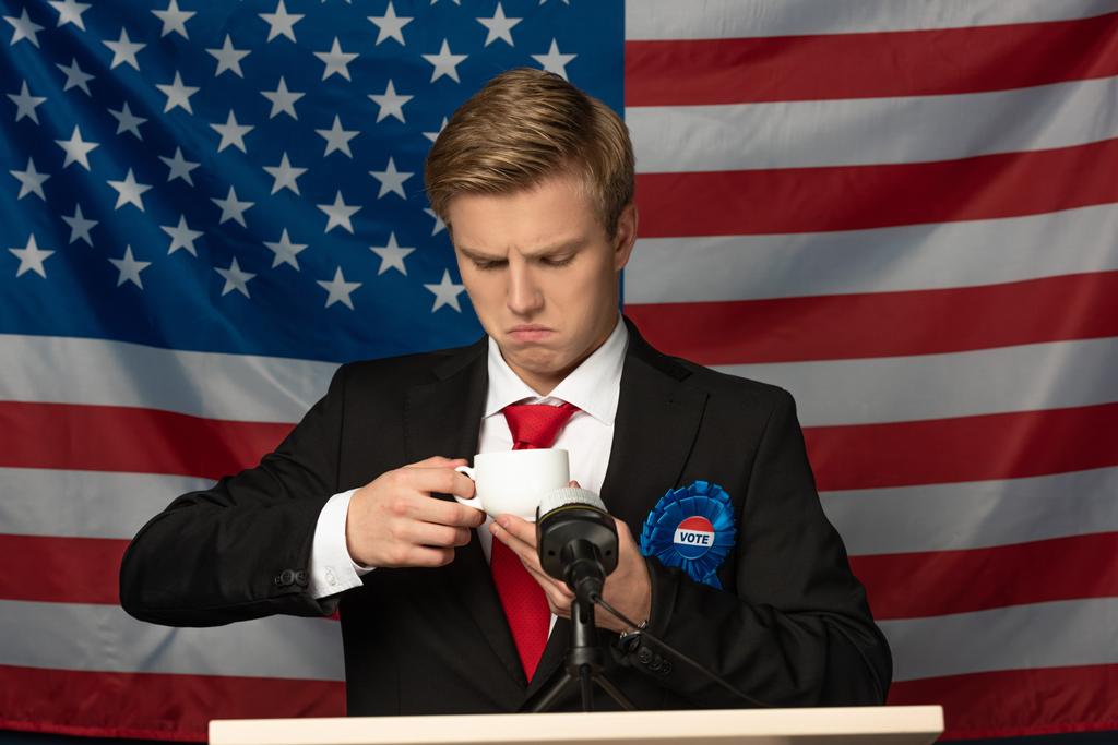 Mies katselee kahvikuppia tribuunissa Amerikan lipun taustalla
 - Valokuva, kuva