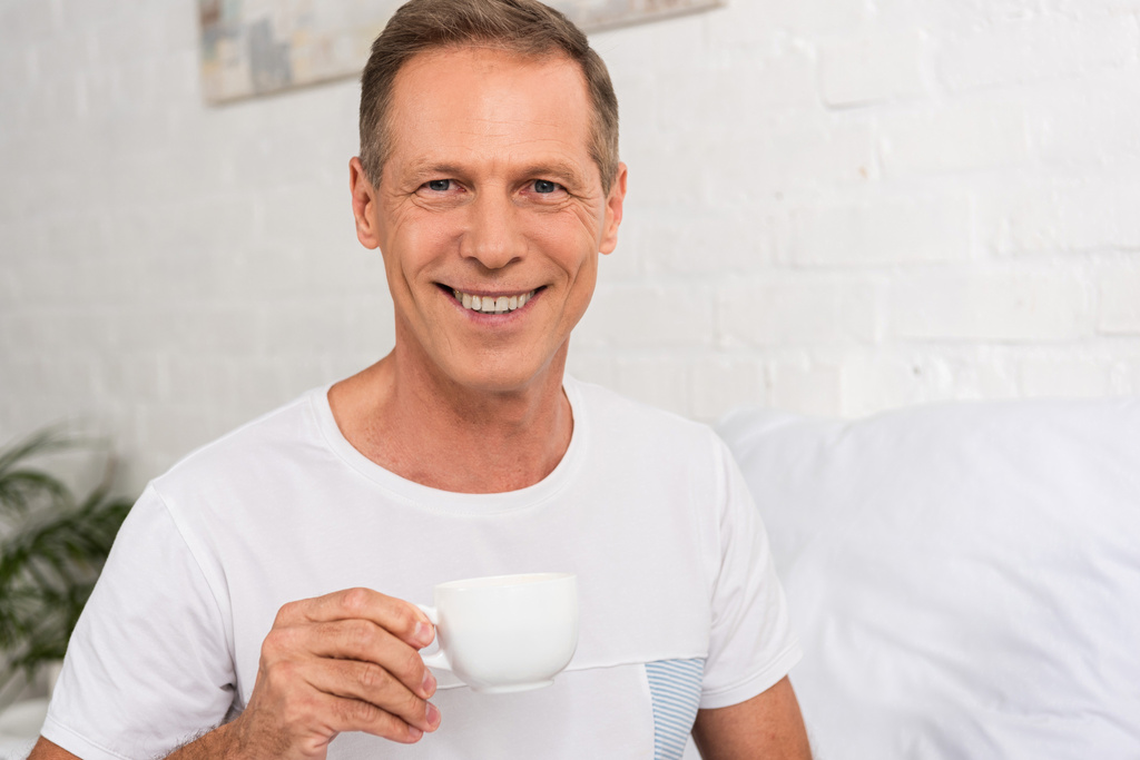 Muž drží šálek kávy a usmívá se na kameru v ložnici - Fotografie, Obrázek