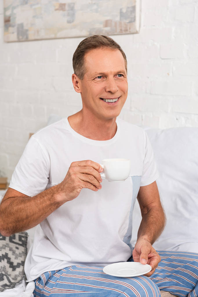 Gülümseyen adam yatakta otururken kahve içiyor. - Fotoğraf, Görsel