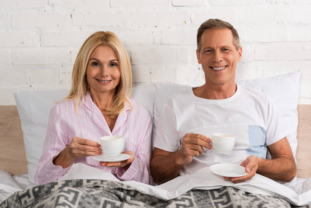 Starší pár s šálky kávy s úsměvem na kameru v posteli - Fotografie, Obrázek