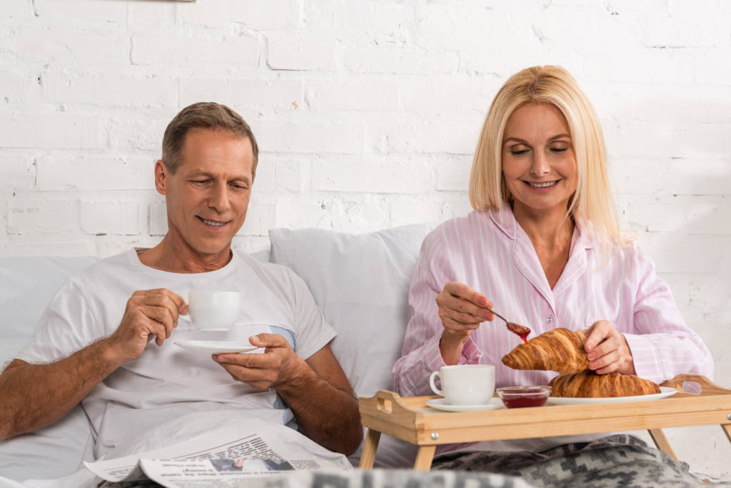 Улыбающаяся пара завтракает, а мужчина читает газету в постели
 - Фото, изображение