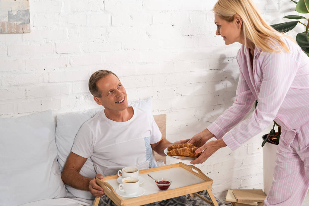 Nainen antaa croissanteja aviomiehelle aamiaistarjottimella sängyssä
 - Valokuva, kuva