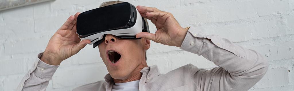 Шокированный человек в наушниках виртуальной реальности, играющий в видеоигру, панорамный снимок
 - Фото, изображение