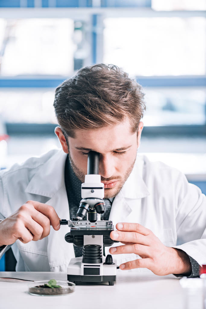 selectieve focus van bebaarde biochemicus in witte vacht kijkend door microscoop in laboratorium  - Foto, afbeelding