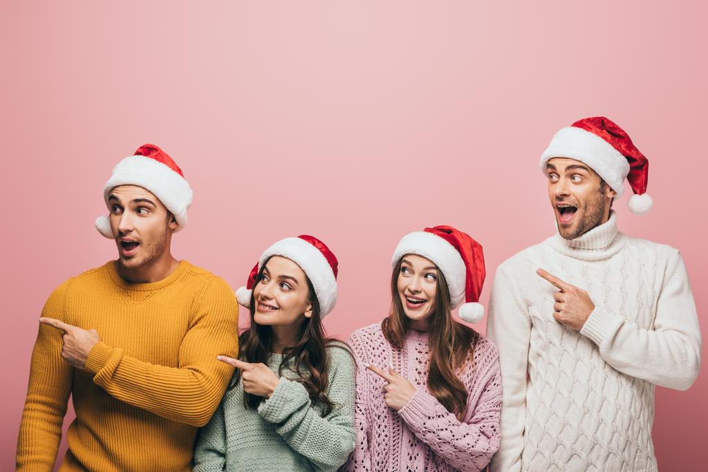 glückliche Freunde in Pullovern und Weihnachtsmützen, die wegzeigen, isoliert auf rosa - Foto, Bild