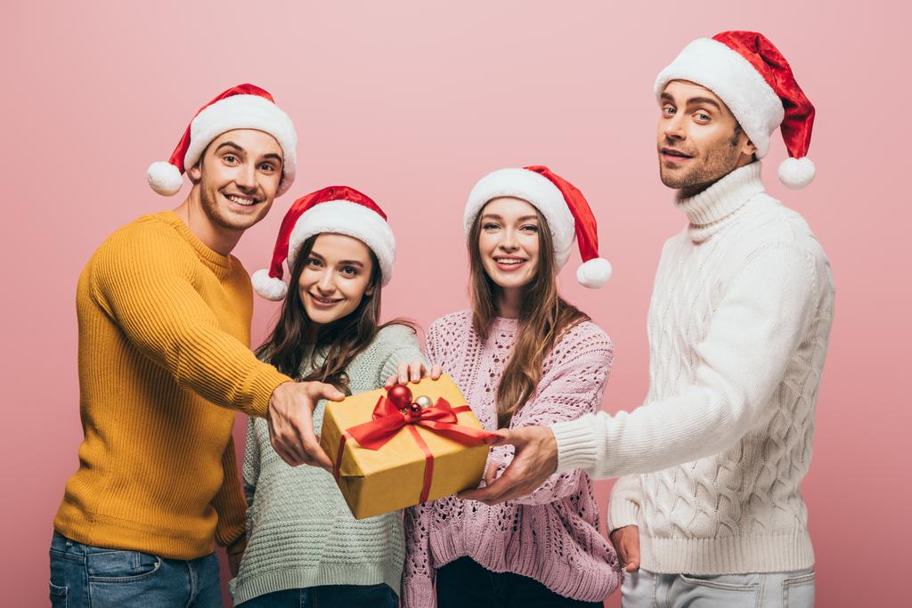 Ευτυχισμένοι φίλοι με καπέλα Σάντα δώρο χριστουγεννιάτικο κουτί δώρου σε σας, απομονώνονται σε ροζ - Φωτογραφία, εικόνα