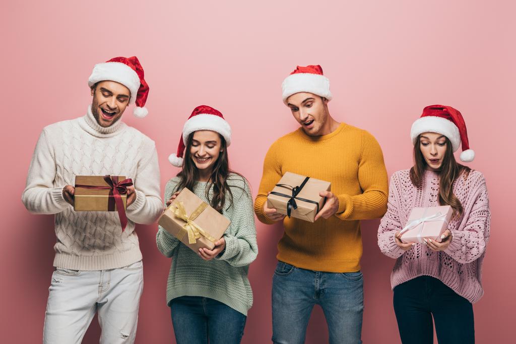 schöne überraschte Freunde in Weihnachtsmützen mit Weihnachtsgeschenken, isoliert auf rosa - Foto, Bild