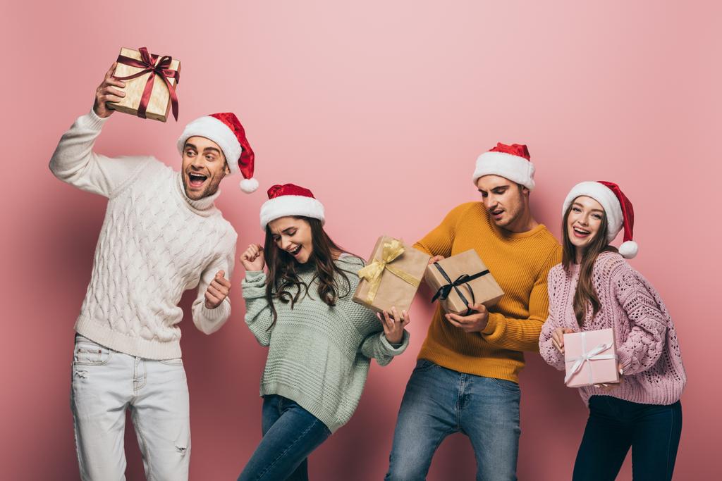 happy friends in santa hats dancing and holding christmas presents, isolated on pink - Photo, Image