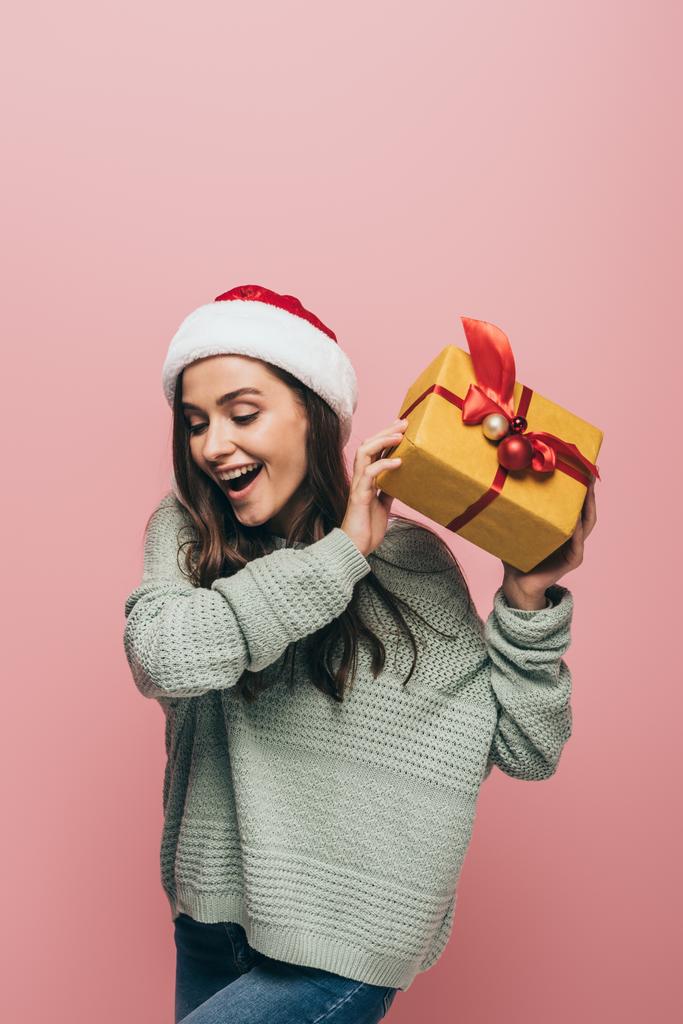 excited woman in sweater and santa hat holding christmas gift, isolated on pink - Photo, Image
