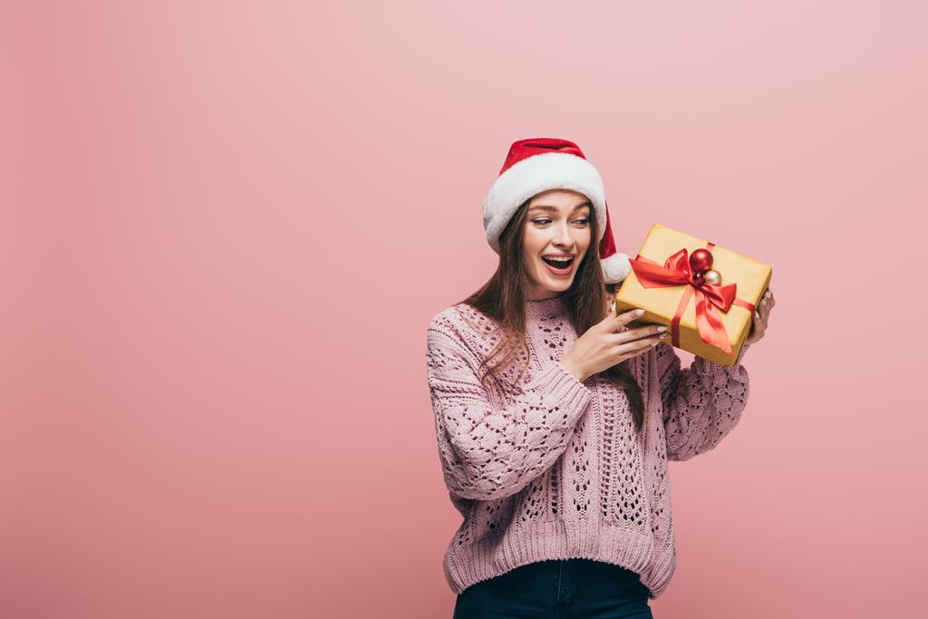 vzrušená žena ve svetru a Santa klobouk drží vánoční dárek, izolované na růžové - Fotografie, Obrázek
