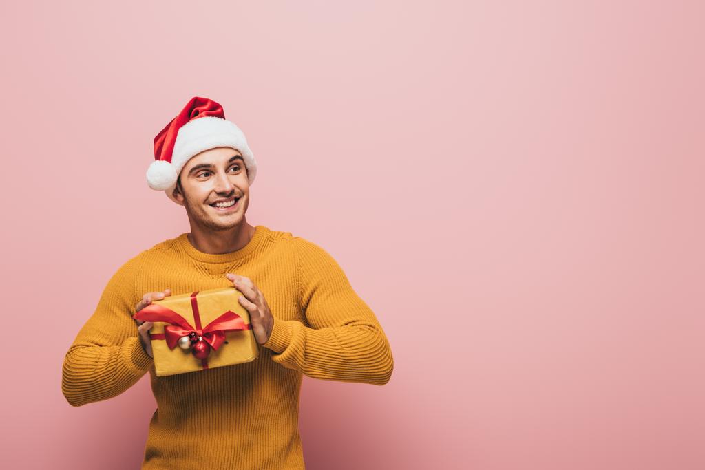mosolygós férfi pulóverben és Mikulás kalap kezében karácsonyi ajándék doboz, elszigetelt rózsaszín - Fotó, kép