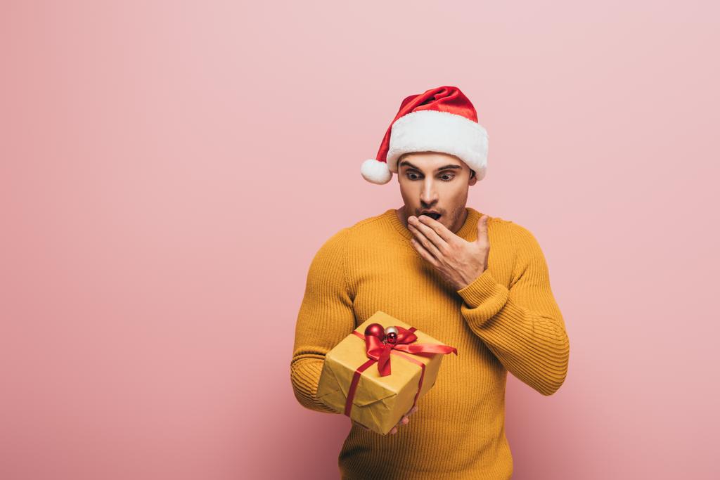 σοκαρισμένος άνθρωπος με πουλόβερ και καπέλο Σάντα κοιτάζοντας το δώρο Χριστουγέννων, απομονώνονται σε ροζ - Φωτογραφία, εικόνα