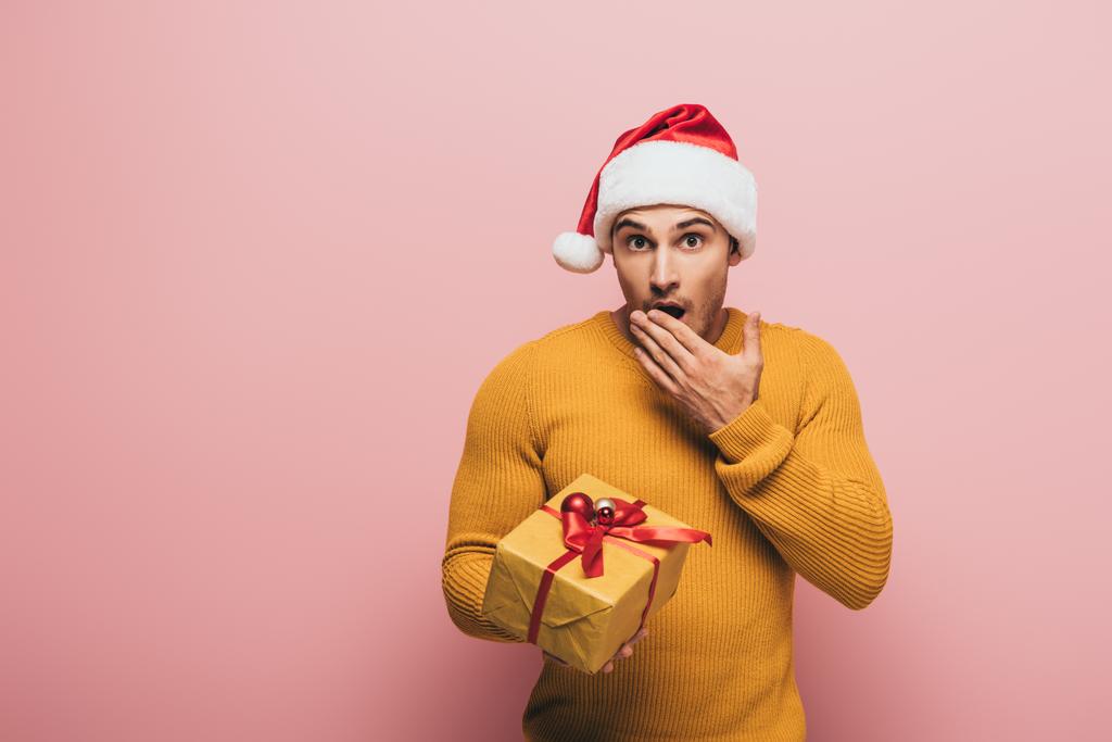 σοκαρισμένος άνθρωπος με το καπέλο santa gesturing και κρατώντας χριστουγεννιάτικο δώρο, απομονώνονται σε ροζ - Φωτογραφία, εικόνα