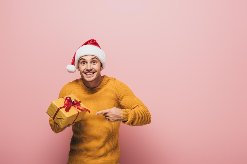 wesoły mężczyzna w swetrze i kapeluszu Mikołaja wskazując na prezent świąteczny, odizolowany na różowy - Zdjęcie, obraz