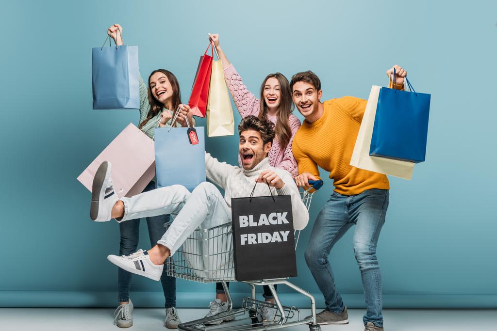 aufgeregte Freunde, die sich am Black Friday mit Einkaufstüten im Warenkorb amüsieren, auf blau - Foto, Bild