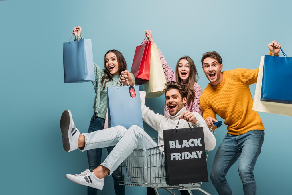 vzrušené mladé přátele baví s nákupními taškami v nákupním košíku v černý pátek, izolované na modré - Fotografie, Obrázek