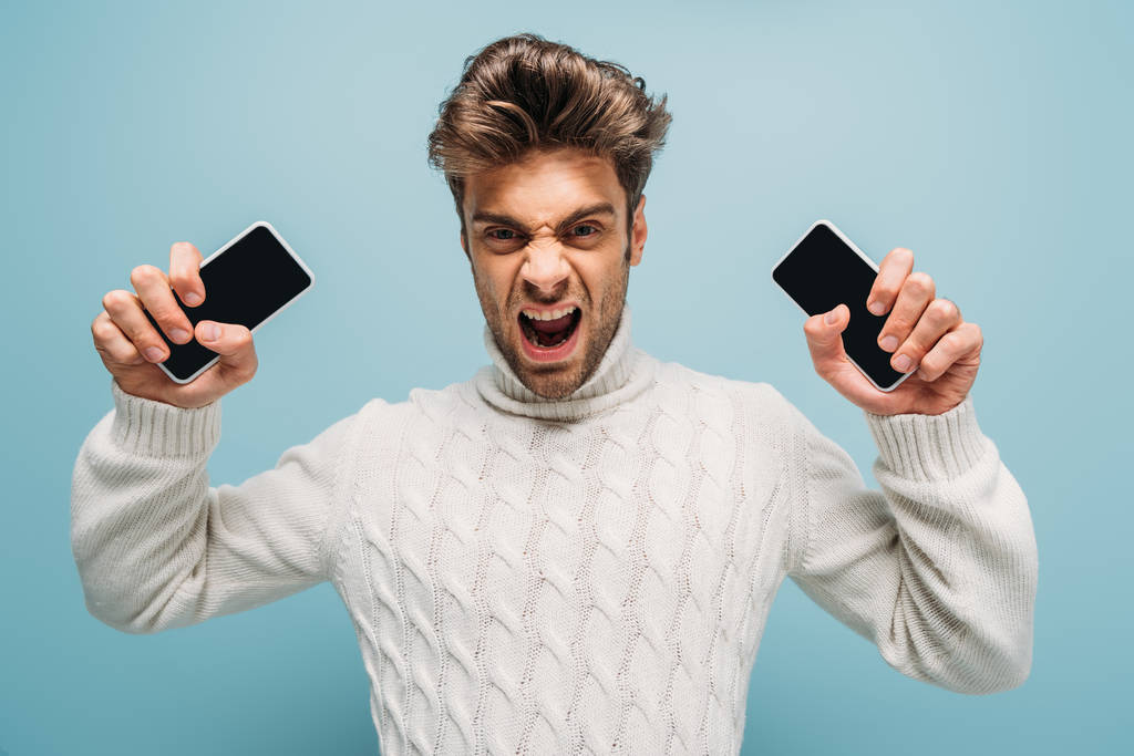 agresivo hombre gritando y sosteniendo dos teléfonos inteligentes con pantallas en blanco, aislado en azul
 - Foto, Imagen