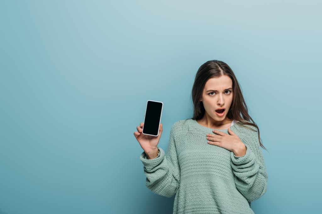 шокована жінка, що показує смартфон з порожнім екраном, ізольована на синьому
 - Фото, зображення
