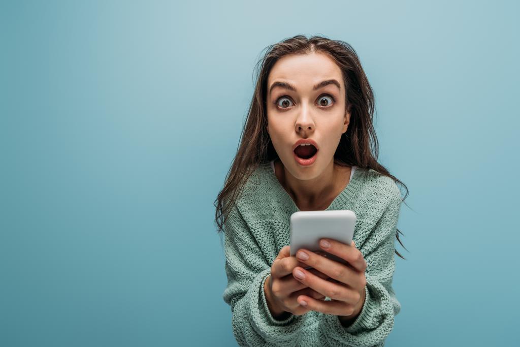 σοκαρισμένη γυναίκα με το στόμα ανοιχτό χρησιμοποιώντας smartphone, απομονώνονται σε μπλε - Φωτογραφία, εικόνα