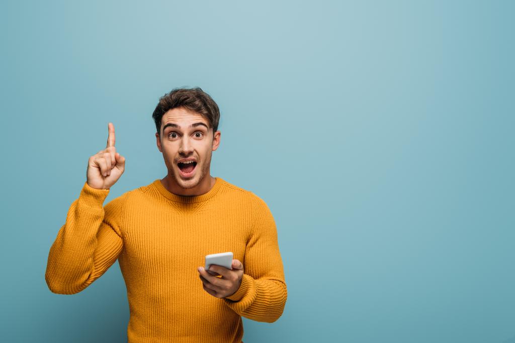 surprised man using smartphone and pointing up, isolated on blue - Photo, Image