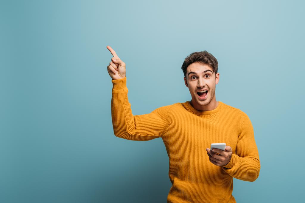 ενθουσιασμένος όμορφος άντρας που δείχνει και χρησιμοποιεί smartphone, απομονωμένος στο μπλε - Φωτογραφία, εικόνα