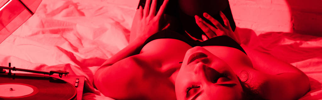 panoramatický záběr atraktivní vášnivé dívky ležící na posteli s vinylovým hráčem v červeném světle - Fotografie, Obrázek