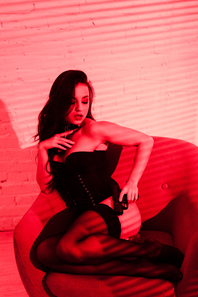 sexy zločinec žena v bodysuit držení zbraň a sedí v křesle na červené - Fotografie, Obrázek