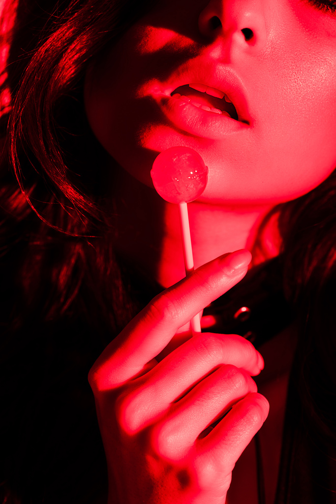 bijgesneden uitzicht van aantrekkelijke gepassioneerde vrouw eten lolly in rood licht - Foto, afbeelding