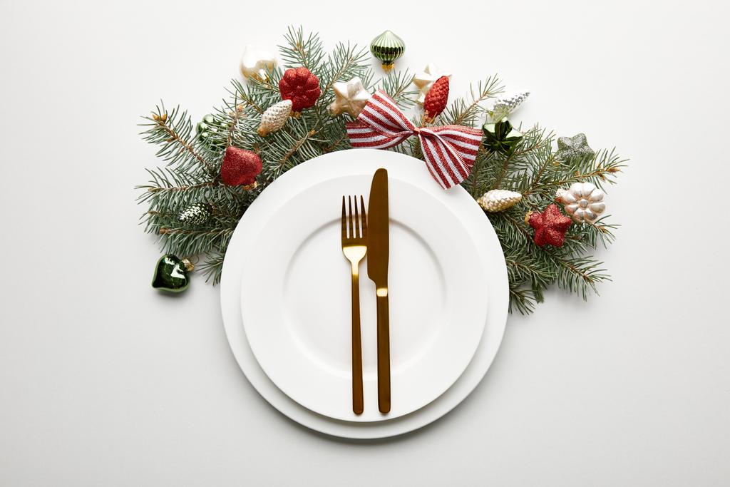 верхний вид на белые тарелки со столовыми приборами рядом с праздничной веткой елки с безделушками на белом фоне
 - Фото, изображение