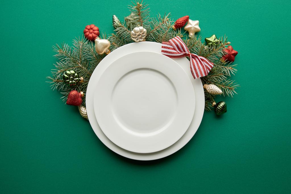верхний вид белых тарелок возле праздничной ветки елки с безделушками на зеленом фоне
 - Фото, изображение