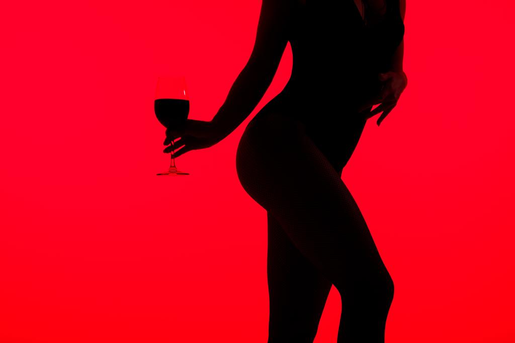 przycięty widok czarnej sylwetki dziewczyny trzymającej kieliszek wina odizolowany na czerwono  - Zdjęcie, obraz