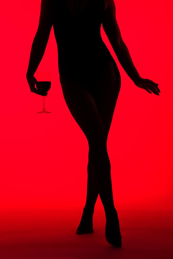 обрезанный вид черного силуэта сексуальной женщины с бокалом вина, изолированного на красный
 - Фото, изображение