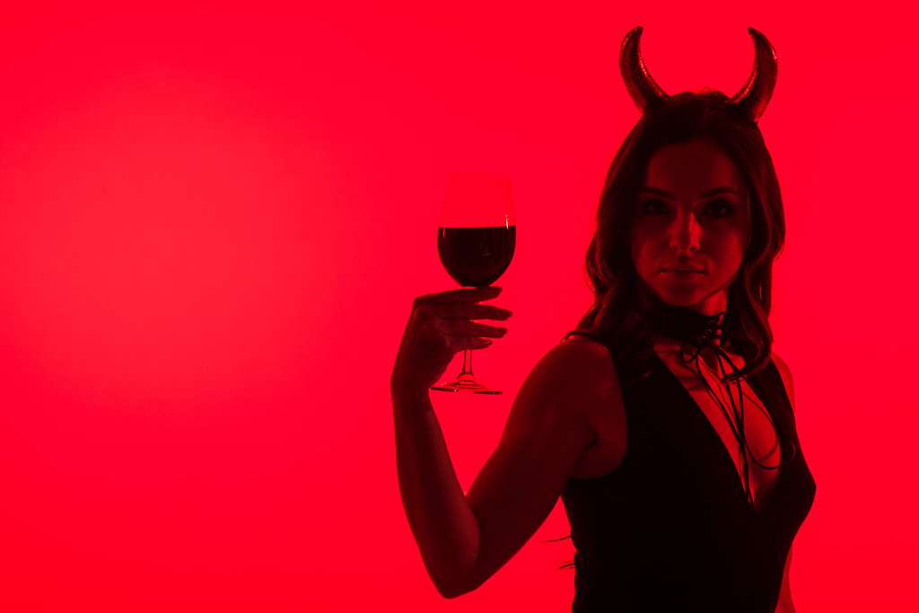 silhouette of sexy girl in Devil costume holding glass of wine, isolated on red - Photo, Image