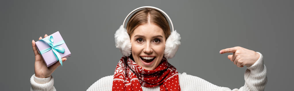панорамный снимок возбужденной девушки в шарфе и подогревателях для ушей, указывающих на рождественский подарок, изолированный на сером
 - Фото, изображение