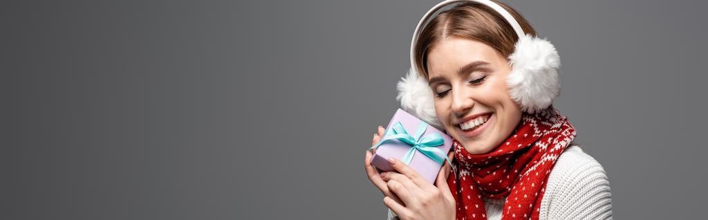 panoramisch schot van gelukkig meisje in sjaal en oorwarmers met kerstcadeau, geïsoleerd op grijs - Foto, afbeelding