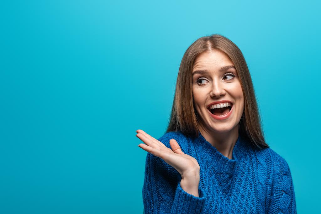 attractive smiling woman with shrug gesture in blue knitted sweater, isolated on blue - Photo, Image