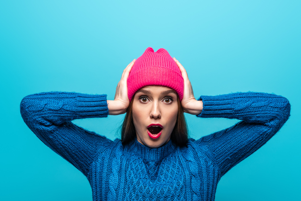 приваблива шокована жінка в трикотажному светрі і рожевому капелюсі, ізольована на синьому
 - Фото, зображення