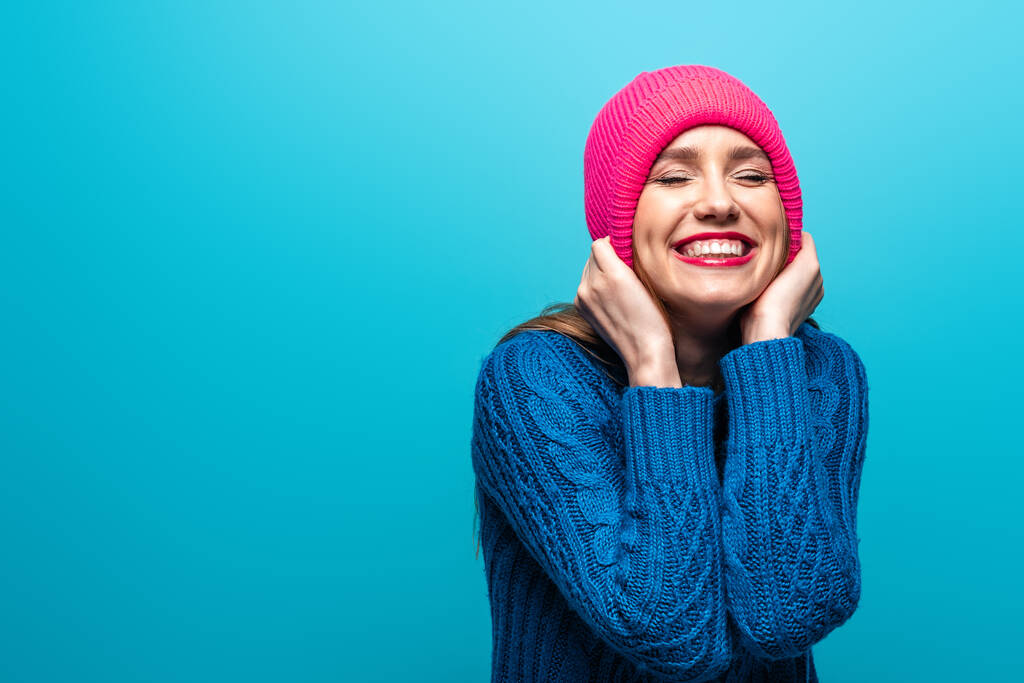 attraktive positive Frau mit geschlossenen Augen in Strickpullover und rosa Mütze, isoliert auf blau - Foto, Bild