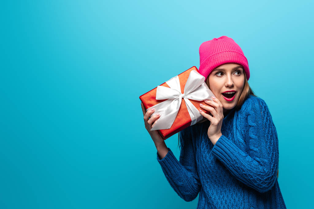 excited girl in knitted sweater and pink hat holding gift box, isolated on blue - Photo, Image