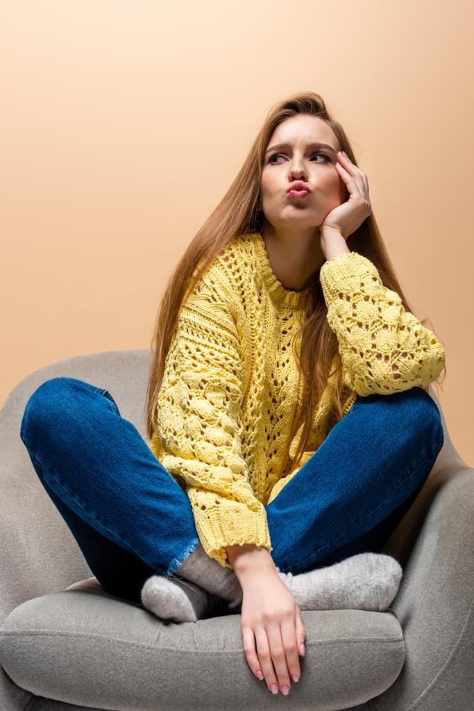 atraente mulher pensativa em suéter amarelo sentado em poltrona, isolado no bege
 - Foto, Imagem