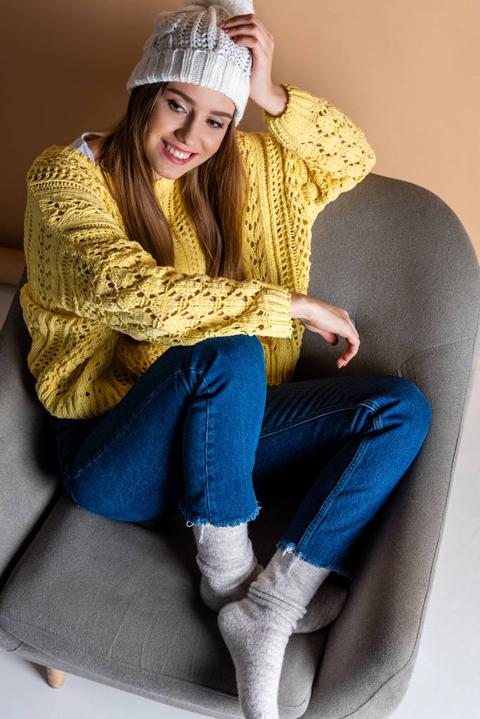 vidám nő sárga pulóverben és kalapban ül karosszékben bézsen - Fotó, kép