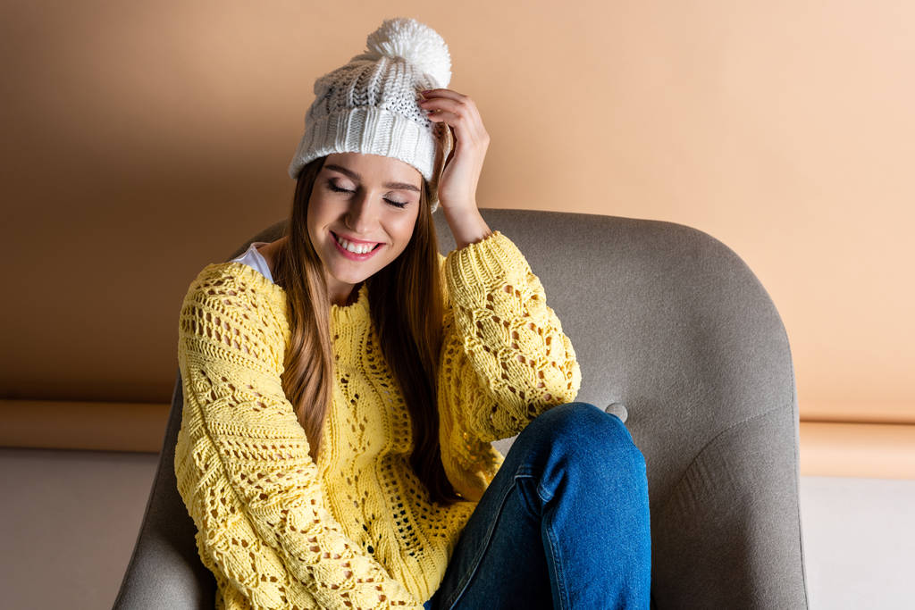 mulher feliz atraente com olhos fechados em suéter amarelo e chapéu sentado em poltrona no bege
 - Foto, Imagem