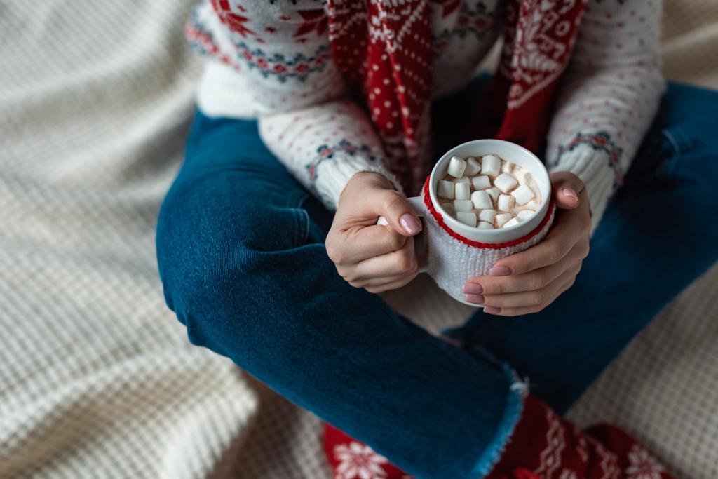 abgeschnittene Ansicht einer Frau im warmen Pullover, die eine Tasse Kakao mit Marshmallow hält und auf einer Decke sitzt - Foto, Bild