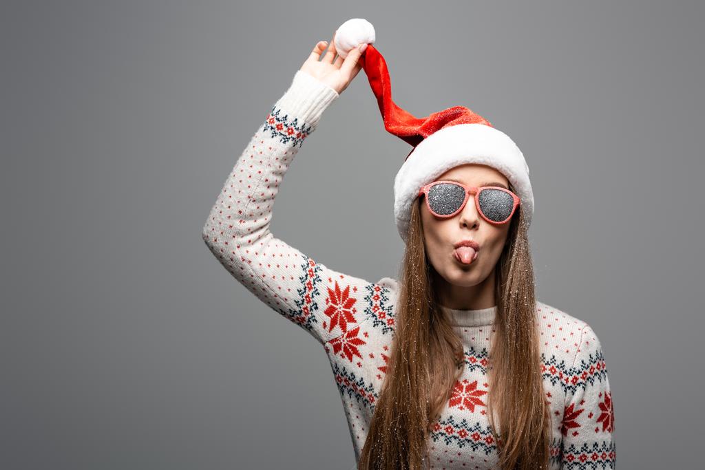 piękna kobieta w świątecznym swetrze, czapce Mikołaja i okularach przeciwsłonecznych wystający język, odizolowana na szaro - Zdjęcie, obraz
