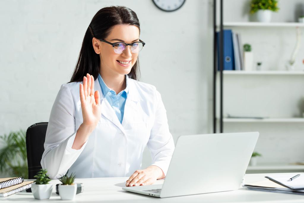 Lächelnder Arzt winkt und hat Online-Konsultation mit Patient auf Laptop in Klinik-Büro - Foto, Bild