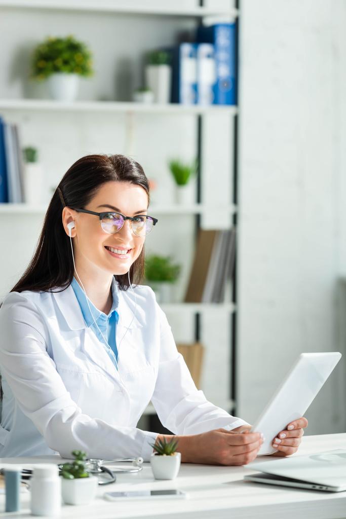 Positiver Arzt mit Online-Beratung auf digitalem Tablet in Klinik-Büro mit Laptop - Foto, Bild