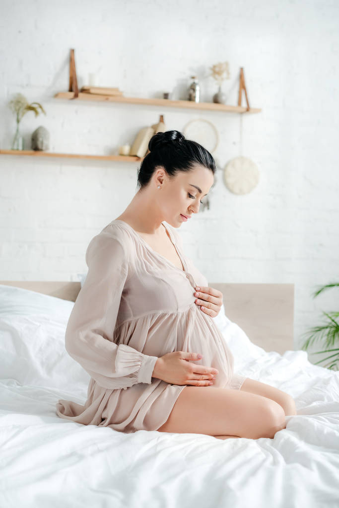 schöne zarte schwangere Frau im Nachthemd berührt Bauch, während sie auf dem Bett sitzt - Foto, Bild