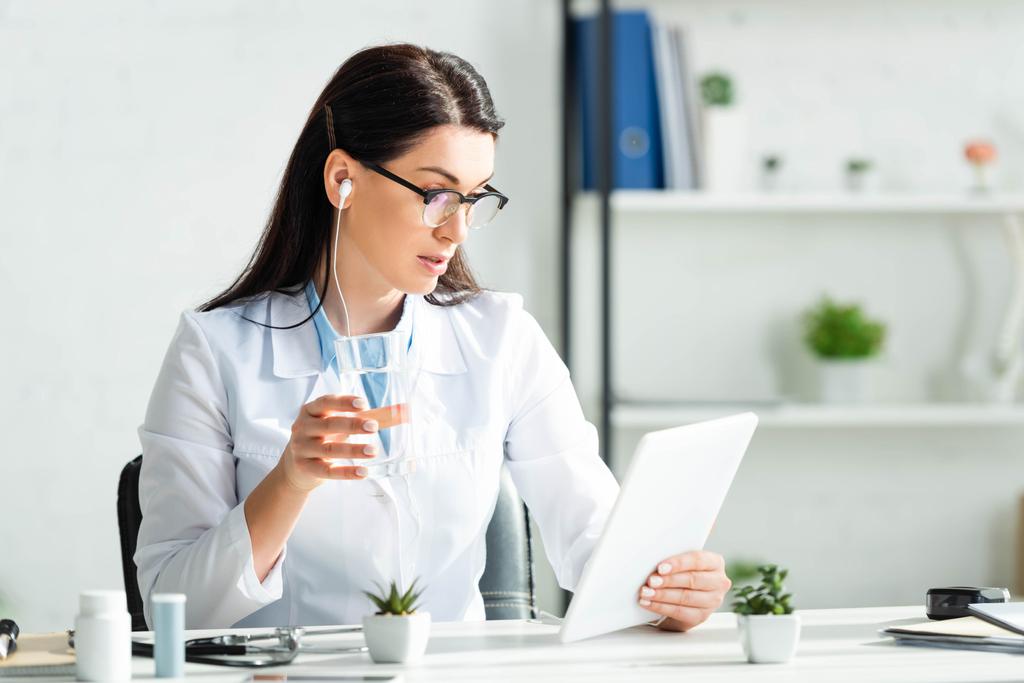 όμορφη γιατρός στα ακουστικά κρατώντας ένα ποτήρι νερό, ενώ έχοντας online διαβούλευση για την ψηφιακή ταμπλέτα στο γραφείο - Φωτογραφία, εικόνα