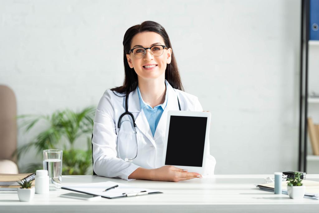 atractivo médico sonriente mostrando tableta digital con pantalla en blanco en la oficina de la clínica
 - Foto, imagen