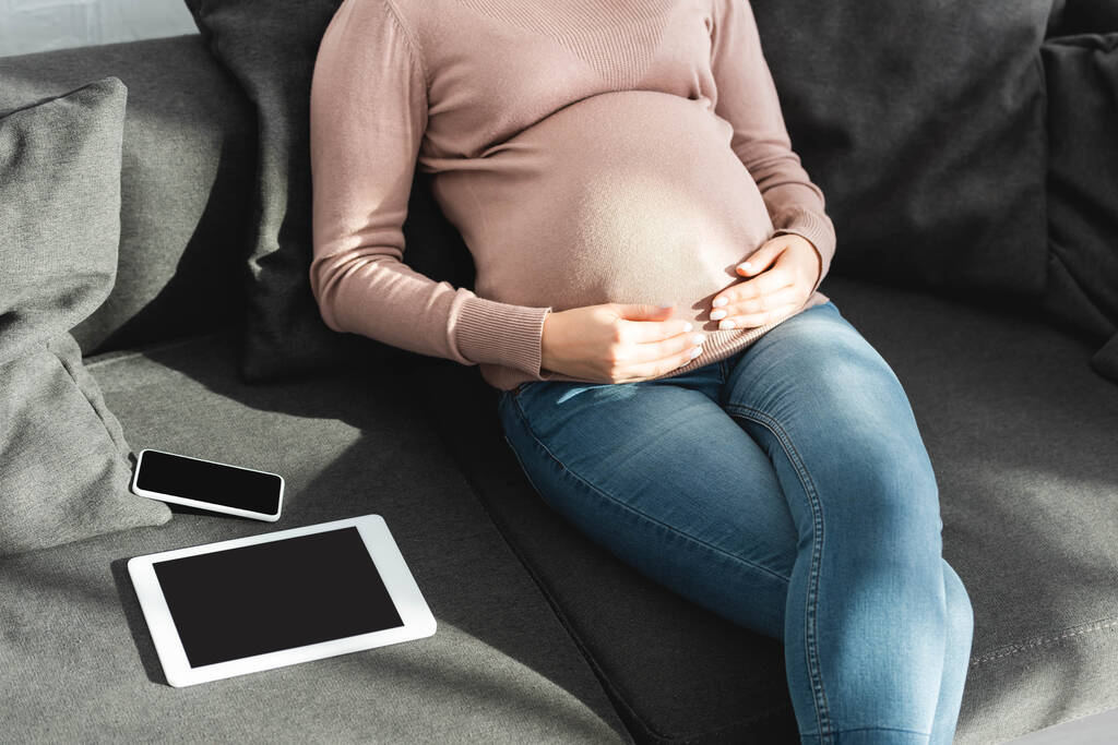 vista recortada de la mujer embarazada tocando el vientre mientras se sienta en el sofá con la tableta digital y el teléfono inteligente
 - Foto, imagen